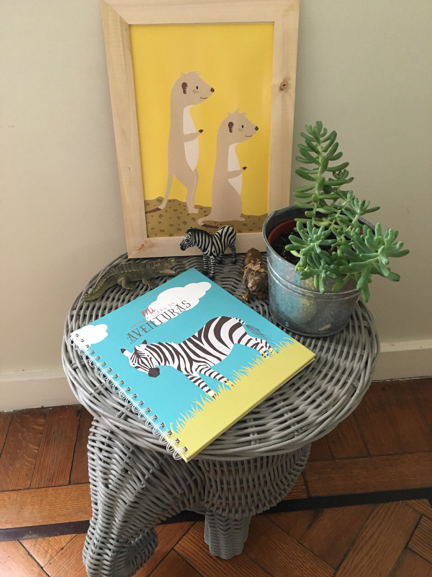 Vista frontal de libro sobre mesa con una planta y poster de suricatas. Fondo de una pared con piso de madera.
